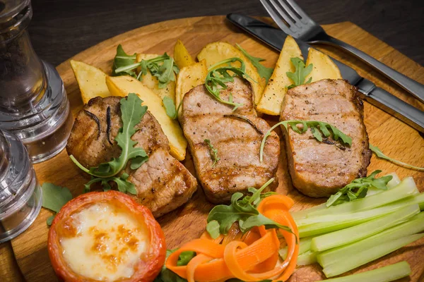 Three pork steaks with fried potatoes and vegetables on a wooden background — Stock Photo, Image
