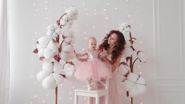 Mooie moeder met een 1-jarige dochter in een fotostudio in de buurt van kunstkatoen en een lichtgevende muur. — Stockvideo