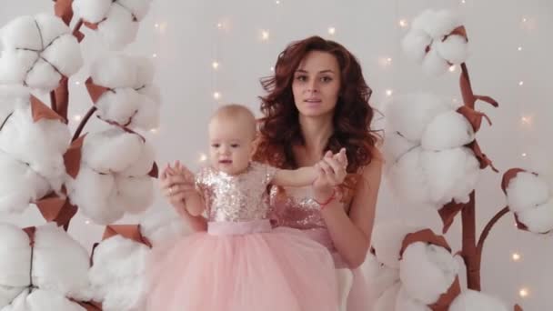 Hermosa madre con una hija de un año en un estudio fotográfico cerca de algodón artificial y una pared luminosa . — Vídeos de Stock