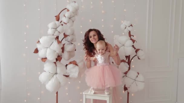 Hermosa madre con una hija de un año en un estudio fotográfico cerca de algodón artificial y una pared luminosa . — Vídeos de Stock