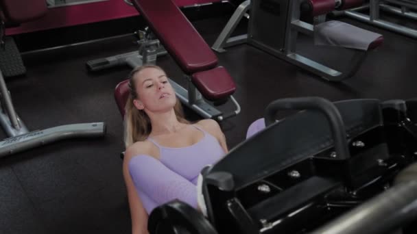 Femme sportive avec une belle silhouette entraîne les jambes sur le simulateur dans la salle de gym . — Video