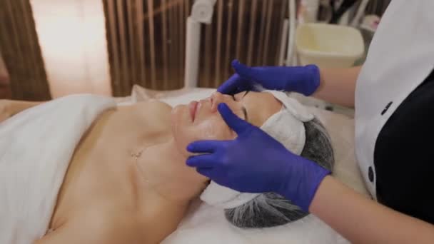 Esteticista profissional aplica óleo de esfoliação a um rosto de mulher com movimentos de massagem das mãos . — Vídeo de Stock