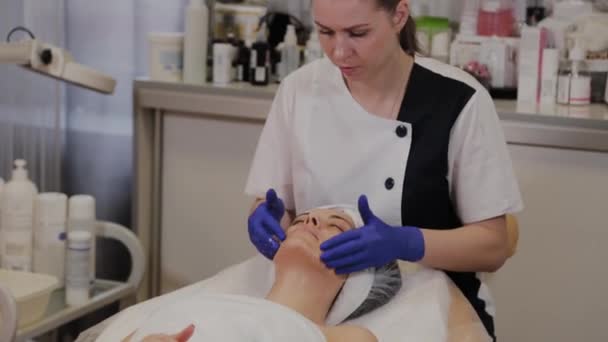 Professionelle Kosmetikerin trägt Peeling-Öl auf ein Frauengesicht mit Massagebewegungen ihrer Hände auf. — Stockvideo