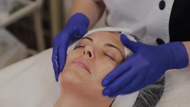 Esteticista profesional aplica aceite exfoliante a una cara de mujer con movimientos de masaje de sus manos . — Vídeos de Stock