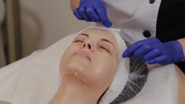 Een professionele schoonheidsspecialiste verwijdert een transparant stof anti-aging masker van een vrouwengezicht. — Stockvideo