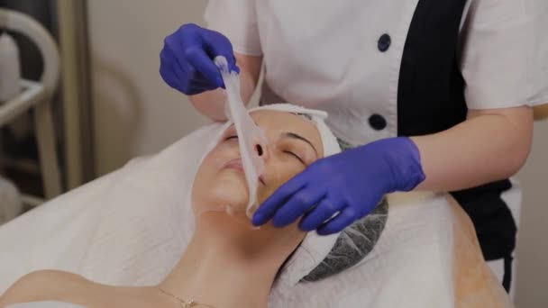 Um esteticista profissional remove uma máscara antienvelhecimento de tecido transparente de um rosto de mulher . — Vídeo de Stock