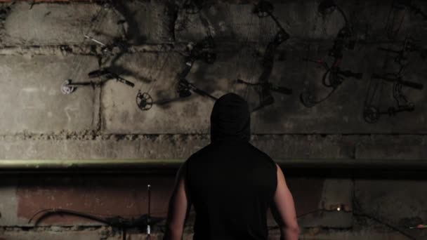 Чоловік в чорному одязі дивиться на стіну з луками і арбалетами . — стокове відео