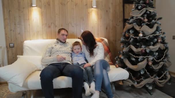 Счастливая семья из трех человек на диване улыбается и разговаривает у елки в рождественскую ночь . — стоковое видео