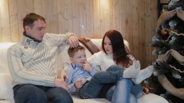 Beautiful happy family of three on the sofa smiling and talking by the Christmas tree on Christmas night. — Stock Video