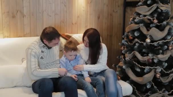 Beautiful happy family of three on the sofa smiling and talking by the Christmas tree on Christmas night. — Stock Video