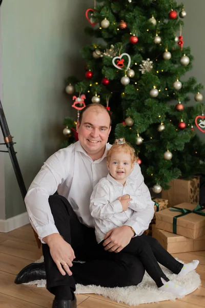 Ein glücklicher Vater mit einer kleinen Tochter im Arm am Weihnachtsbaum. Frohe Weihnachten 2020. — Stockfoto