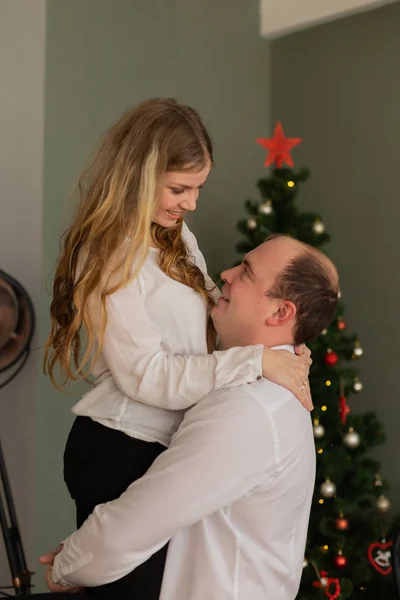 Šťastný manžel a žena na vánočním stromečku doma. Manžel drží svou ženu v náručí. — Stock fotografie