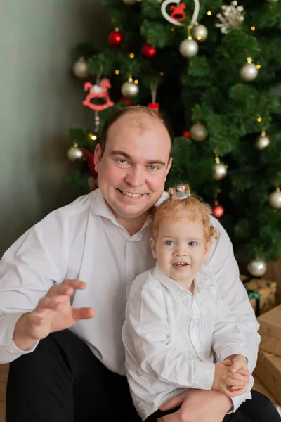 Ein glücklicher Vater mit einer kleinen Tochter im Arm am Weihnachtsbaum. Frohe Weihnachten 2020. — Stockfoto