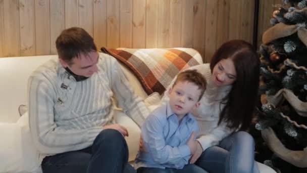 Belle famille heureuse de trois personnes sur le canapé souriant et parlant près du sapin de Noël la nuit de Noël . — Video