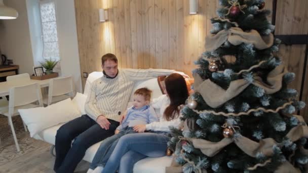 Hermosa familia feliz de tres en el sofá sonriendo y hablando junto al árbol de Navidad en la noche de Navidad . — Vídeo de stock