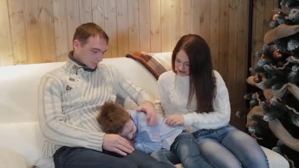 Belle famille heureuse de trois personnes sur le canapé souriant et parlant près du sapin de Noël la nuit de Noël . — Video