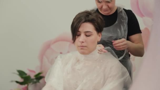 Professional hairdresser woman putting cape on girl. — Stock Video