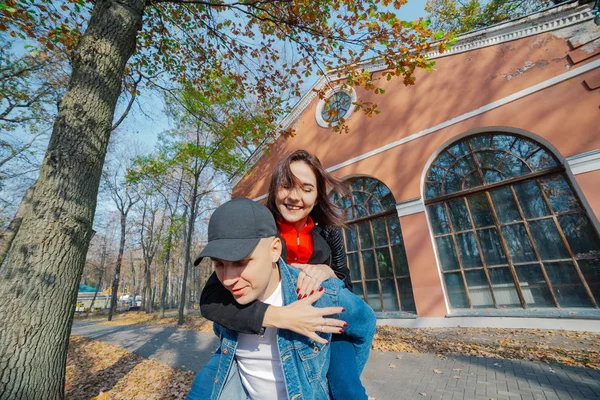 Young beautiful couple and their labrador in the Park. A man holds a woman on his back and she hugs him. Rays of setting sun break through the branches of trees. Vertically framed shot.. — Stock Photo, Image