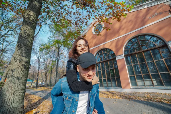 Junges schönes Paar und ihr Labrador im Park. Ein Mann hält eine Frau auf dem Rücken und sie umarmt ihn. Sonnenstrahlen brechen durch die Äste der Bäume. vertikal gerahmte Aufnahme.. — Stockfoto