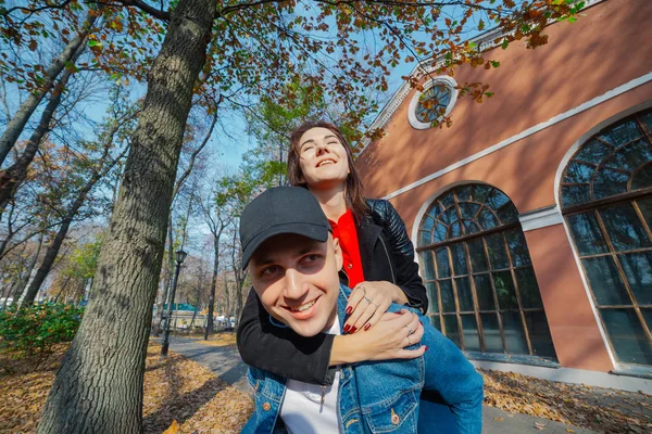 Young beautiful couple and their labrador in the Park. A man holds a woman on his back and she hugs him. Rays of setting sun break through the branches of trees. Vertically framed shot.. — Stock Photo, Image