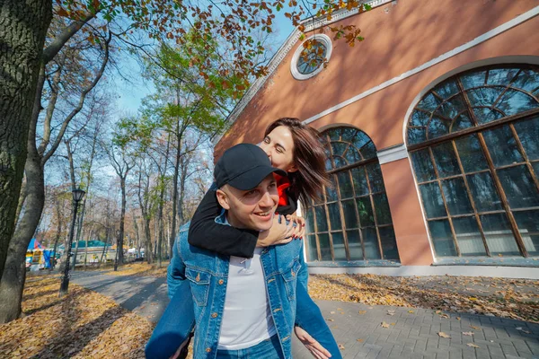 Joven hermosa pareja y su labrador en el Parque. Un hombre sostiene a una mujer en su espalda y ella lo abraza. Los rayos del sol poniente rompen las ramas de los árboles. Tiro enmarcado verticalmente .. —  Fotos de Stock