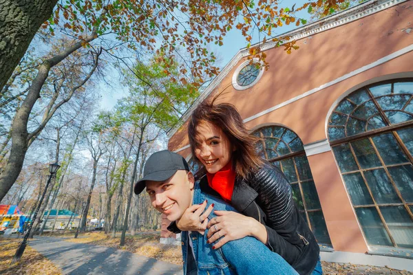 Young beautiful couple and their labrador in the Park. A man holds a woman on his back and she hugs him. Rays of setting sun break through the branches of trees. Vertically framed shot.. — Stock Photo, Image