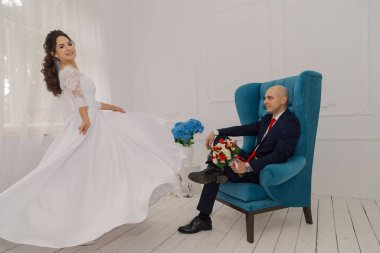 Yakışıklı bir damat sandalyede oturuyor. Gelin ona elbisesini gösterir..