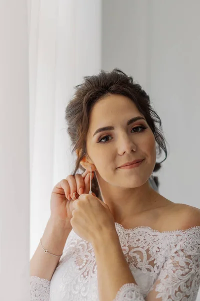 Beautiful bride puts on earrings by the window. Happy wedding day. — Stock Photo, Image