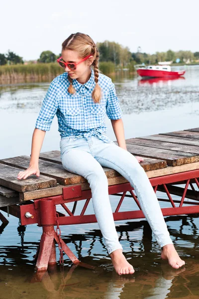 Young girl sitting on jetty over the lake and dipping feet in wa — Stock Photo, Image