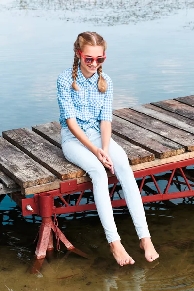 Usmívající se dívka sedí na molu nad jezerem a máčení nohou v — Stock fotografie