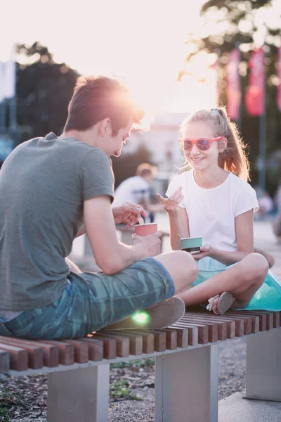 Девочка-подросток и мальчик проводят время вместе в центре города, наслаждаясь мороженым в летний день. Проводить время в солнечный день, поедая сладкий десерт. Центр города на заднем плане — стоковое фото