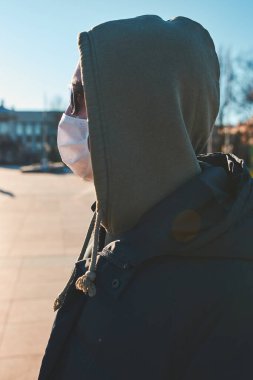 Virüs bulaşmasını önlemek ve hastalığın yayılmasını önlemek için maske takan kapüşonlu genç bir adam. Şehir merkezinde yürüyüş. Virüs enfeksiyonu koruması. Gerçek insanlar, otantik durumlar.