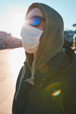 Virüs bulaşmasını önlemek ve hastalığın yayılmasını önlemek için maske takan kapüşonlu genç bir adam. Şehir merkezinde yürüyüş. Virüs enfeksiyonu koruması. Gerçek insanlar, otantik durumlar.