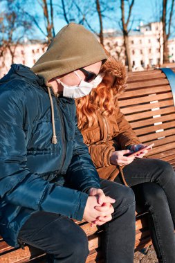 Virüs bulaşmasını önlemek ve hastalığın yayılmasını önlemek için yüz maskesi takan genç adam ve kadın. Şehir merkezindeki bankta oturan çift. Virüs enfeksiyonu koruması. Gerçek insanlar, otantik durumlar.