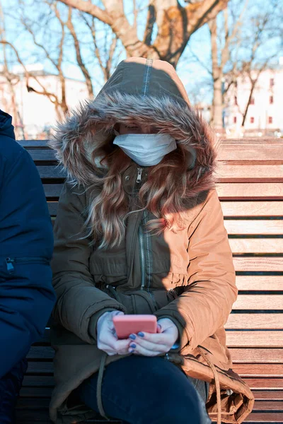Girl wearing the face mask to avoid virus infection and to prevent the spread of disease. Young woman sitting on a bench in city centre. Virus infection protection. Real people, authentic situations