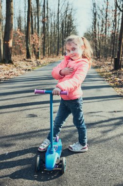Parkta oynayan, scooter süren, güneşli bir sonbahar gününde eğlenen küçük bir kız. Gerçek insanlar, otantik durumlar.