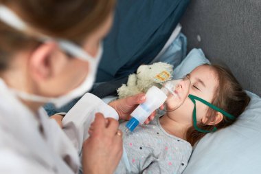 Doktor evde küçük bir hastayı ziyaret ediyor. Çocuk nebulizörle tıbbi solunum tedavisi görüyor. Yüzünde solunum maskesi olan bir kız. Üniformalı ve maskeli bir kadın.