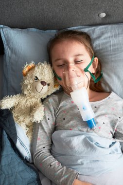 Hasta küçük kız nebuliserle tıbbi solunum tedavisi görüyor. Yüzünde nefes maskesi olan çocuk yatakta yatıyor.