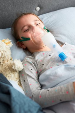 Hasta küçük kız nebuliserle tıbbi solunum tedavisi görüyor. Yüzünde nefes maskesi olan çocuk yatakta yatıyor.