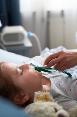Doktor evde küçük bir hastayı ziyaret ediyor. Çocuk nebulizörle tıbbi solunum tedavisi görüyor. Yüzünde solunum maskesi olan bir kız. Üniformalı ve maskeli bir kadın.