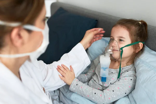Doctor visiting little patient at home. Child having medical inhalation treatment with nebuliser. Girl with breathing mask on her face. Woman wearing uniform and face mask