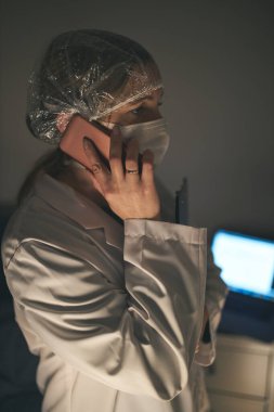Doktor bir telefon görüşmesi yapıyor. Hastane personeli gece vardiyasında çalışıyor. Virüs enfeksiyonunu önlemek için üniforma, şapka ve yüz maskesi takan kadın.