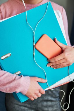 Kadın, COVID-19 karantinası sırasında evden telefonla konuşma dersleri sırasında uzaktan ödev yapmayı öğreniyor. Belgeleri klasörde tutan kadın kulaklık ve akıllı telefon kullanıyor.