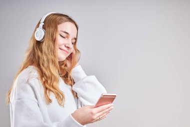 Müzik dinleyen genç bir kız video izlerken eğleniyor arkadaşlarıyla sohbet ediyor akıllı telefon kulaklıkları takıp gri arka planda dikiliyor.