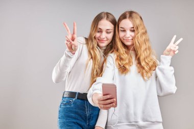 Birlikte müzik dinleyen genç kızlar video izleyerek eğleniyorlar görüntülü sohbet yapıyorlar arkadaşlarla sohbet ediyorlar akıllı telefon kulaklıkları takıp gri arka planda duruyorlar.