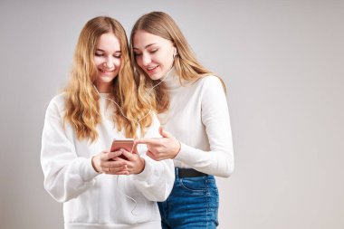 Birlikte müzik dinleyen genç kızlar video izleyerek eğleniyorlar görüntülü sohbet yapıyorlar arkadaşlarla sohbet ediyorlar akıllı telefon kulaklıkları takıp gri arka planda duruyorlar.