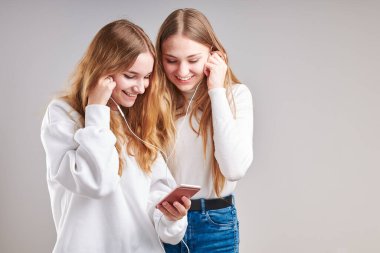 Birlikte müzik dinleyen genç kızlar video izleyerek eğleniyorlar görüntülü sohbet yapıyorlar arkadaşlarla sohbet ediyorlar akıllı telefon kulaklıkları takıp gri arka planda duruyorlar.