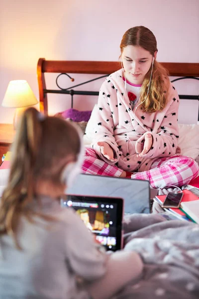 Student learning online, watching lesson remotely, listening to professor, talking with classmates on video call from home during quarantine. Young girl using laptop, headphones, books, manuals sitting in bed