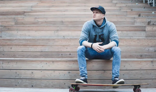 Привлекательный молодой человек сидит на деревянных ступеньках в городском парке со своим скейтбордом — стоковое фото