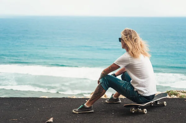 Jovem homem skatista loira sentado na frente da vista incrível do oceano — Fotografia de Stock